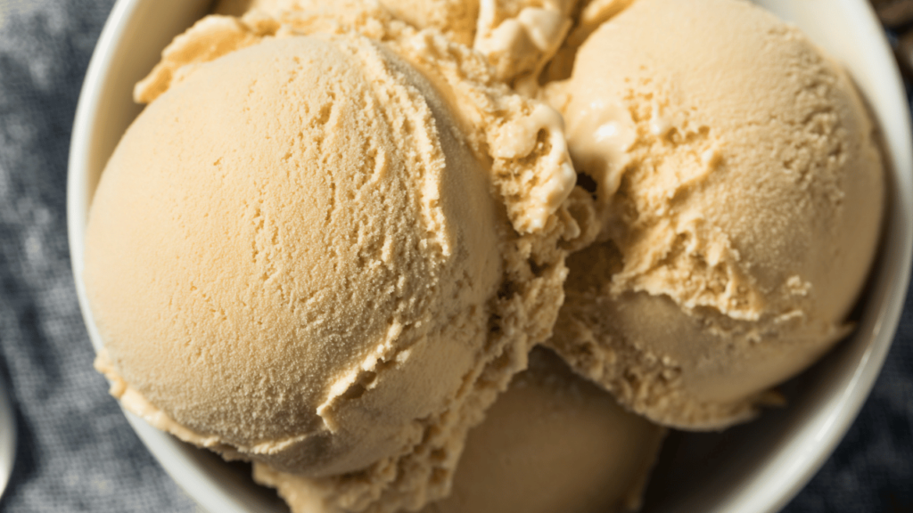 A vibrant kitchen scene showcasing the process of making Butterfinger ice cream. In the foreground, a stainless steel ice cream maker sits on a marble countertop, elegantly churned ice cream in progress, with a scoop of crushed Butterfinger candy beside it. In the middle, various ingredients like a bowl of cream, a jar of sweetened condensed milk, and a handful of Butterfinger pieces are artistically arranged. The background features a cozy kitchen with warm, natural light streaming through a window, subtly illuminating the workspace. Add a soft-focus effect to create a dreamy atmosphere, emphasizing the creamy texture and delightful colors of the ice cream. The overall mood is inviting and cheerful, evoking the joy of homemade treats.