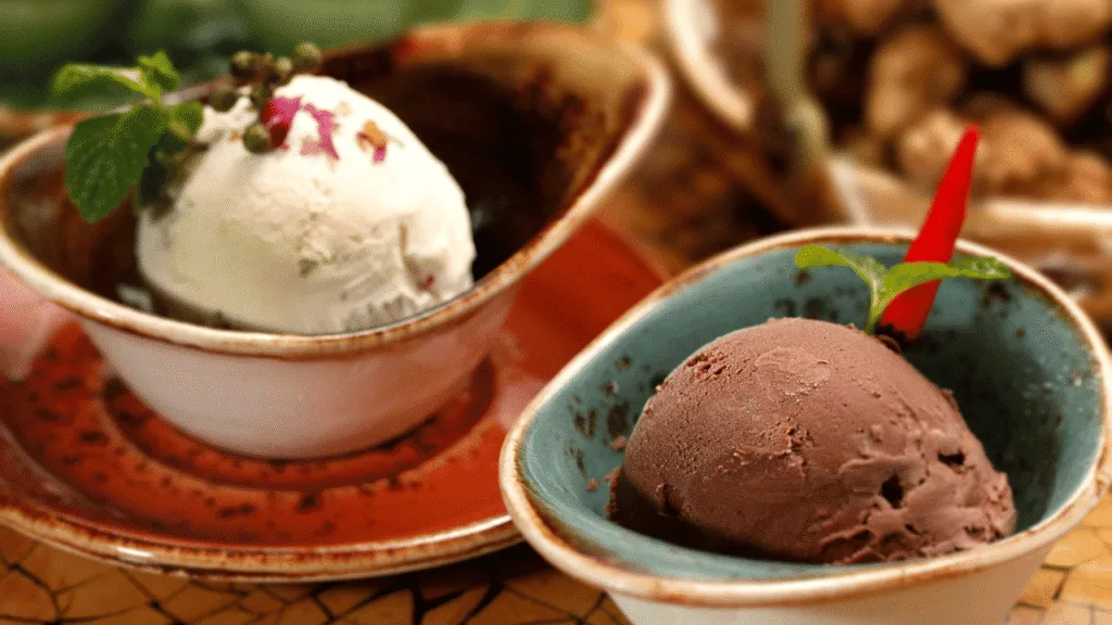 Vivid, brightly lit image of an assortment of unusual vegan ice cream flavors, captured from an overhead angle with a shallow depth of field. In the foreground, a selection of scoops in intriguing hues like matcha green, purple sweet potato, and turmeric-spiced coconut, arranged artfully in a sleek stainless steel or glass container. In the middle ground, complementary toppings and mix-ins such as candied ginger, toasted coconut flakes, and crushed nuts. The background softly blurred, suggesting a modern, minimalist kitchen or cafe setting with clean lines and a light, airy atmosphere. Dairy Free Sugar Free Ice Cream Experts Share Their Favorite Flavor Combinations