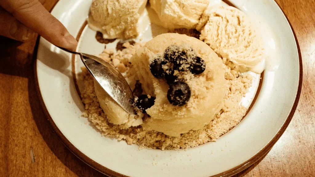 A step-by-step scene showing the process of churning old-fashioned ice cream. In the foreground, a vintage metal ice cream maker sits on a wooden table, its crank handle being turned by a person's hands. Nearby, a bowl of cream and sugar sits ready for mixing. The middle ground reveals the interior mechanism of the churn, with the churning paddle slowly rotating to aerate the mixture. In the background, a rustic kitchen setting with simple wooden shelves, jars, and utensils, all bathed in warm, soft lighting that creates a cozy, nostalgic atmosphere. The focus is on the tactile, manual process of making homemade ice cream, evoking feelings of tradition and timelessness. Old Fashioned Ice Cream Churn Step-by-Step Guide to Churning Ice Cream
