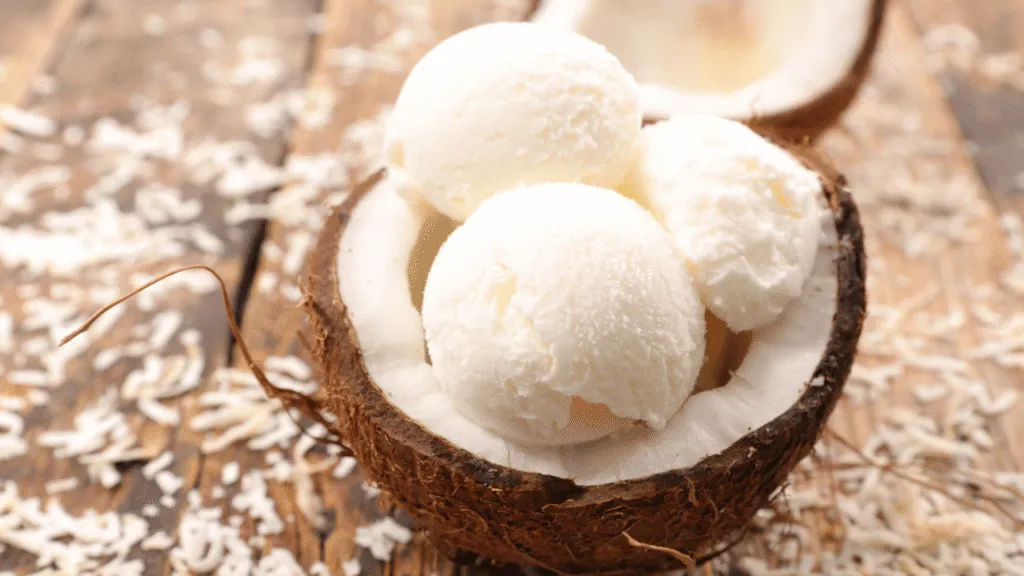 A simply delightful coconut ice cream, with a smooth and creamy texture, sitting atop a bright white ceramic bowl. The frozen dessert glistens under the warm golden light, casting soft shadows across its surface. The bowl is nestled on a wooden table, surrounded by a few fresh coconut halves and a sprig of mint, adding a touch of natural elegance. The overall scene exudes a calming, homemade atmosphere, perfectly capturing the essence of creating this simple yet delicious treat in your own kitchen. Simple Steps to Make Your Coconut Ice Cream Recipe