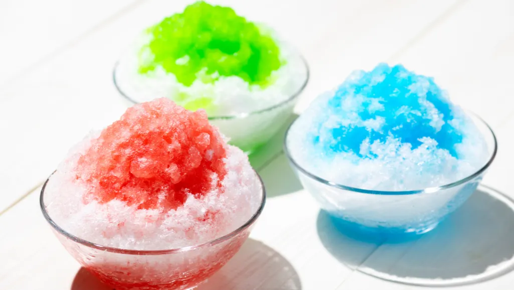 A vintage snow cone machine surrounded by delicate, shaved ice crystals cascading down its sides, creating a refreshing and nostalgic scene. The ice glistens under soft, warm lighting, casting a gentle glow throughout the frame. In the background, a classic pastel-colored tiled floor and a hint of retro-inspired decor set the stage for this timeless treat. The composition emphasizes the tactile nature of the shaved ice, inviting the viewer to imagine the cool, smooth texture and the sweet flavors that might accompany this beloved summertime delight.