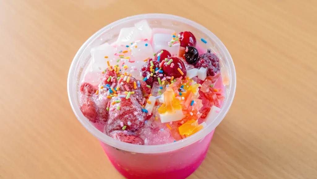 A glass jar filled with a vibrant, jewel-toned homemade snow cone syrup, its surface glistening under warm, natural lighting. The syrup's deep hues of red, blue, and green create a captivating contrast against the clear glass container. Reflected on the surface, you can see a stainless steel spoon, hinting at the delicious and refreshing treat this syrup will become. The jar is placed on a wooden table, with a soft, blurred background that suggests a cozy, homemade setting. The overall mood is one of simplicity, homemade charm, and the promise of a sweet, icy delight.