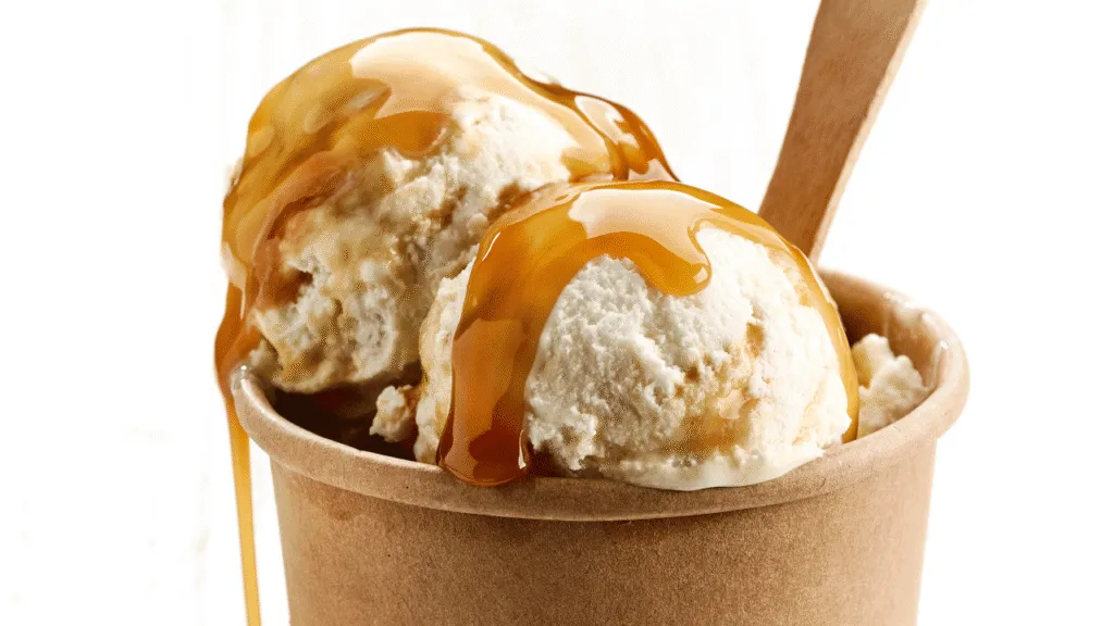 A neatly organized kitchen counter, its surface illuminated by soft, natural lighting. On the counter, a glass bowl filled with a rich, golden-colored butterscotch sauce, slowly being swirled and folded with a whisk, creating mesmerizing patterns and ribbons. The smooth, velvety texture of the sauce is evident, as it gently flows and combines with the motions of the whisk. In the background, a metal saucepan simmers, hinting at the custard base being prepared, essential for the perfect butterscotch ice cream. The overall scene exudes a warm, cozy atmosphere, inviting the viewer to imagine the delightful flavors and textures that will soon come together. Crafting the Butterscotch Sauce and Custard Base