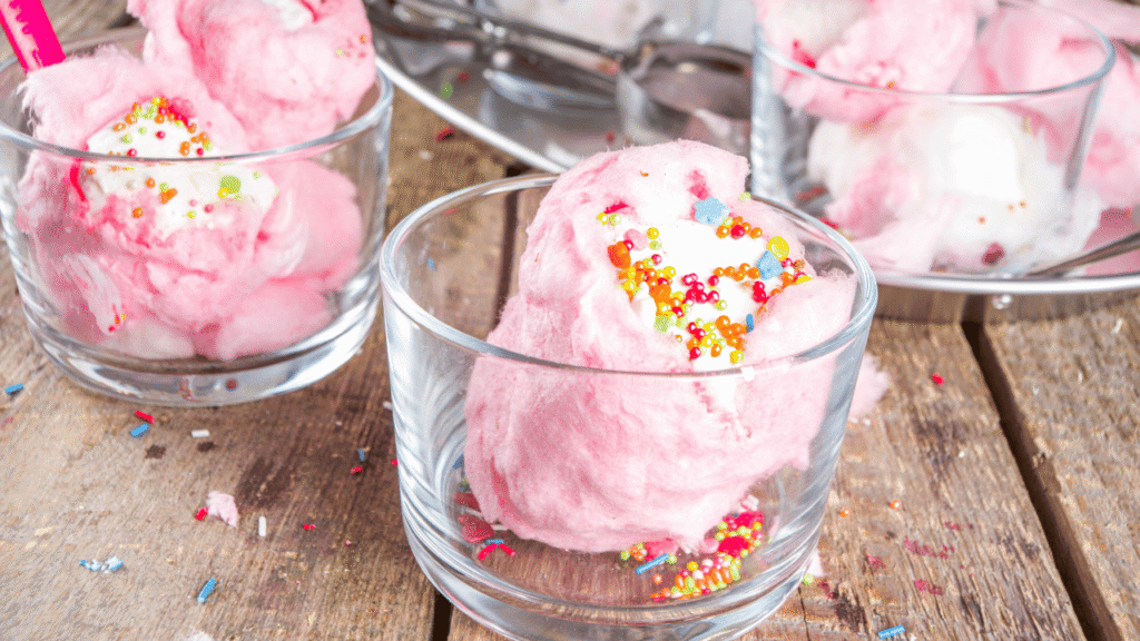A whimsical close-up of cotton candy-inspired ice cream, its pastel pink and blue hues swirling together in a frozen treat. Fluffy, cloud-like scoops glisten under a warm, golden light, casting soft shadows that accentuate the luscious texture. The creamy surface is punctuated by delicate, wispy sugar strands in matching colors, evoking the airy, spun-sugar aesthetic of classic carnival fare. The composition focuses on the dessert's captivating visual appeal, inviting the viewer to indulge in its enchanting, nostalgic flavors. Exploring Cotton Candy Ice Cream Recipe Flavor and Visual Appeal