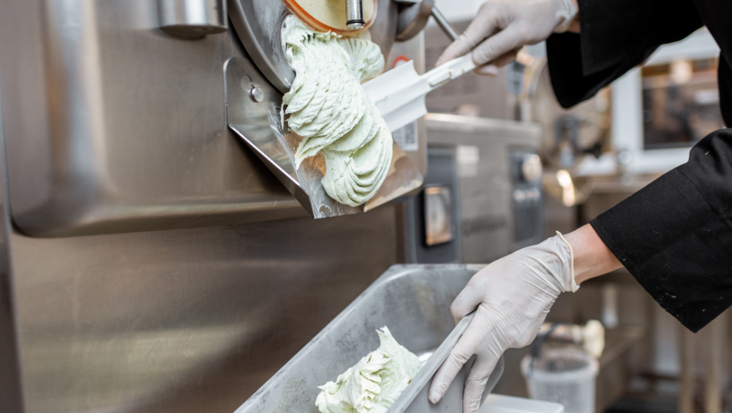 A stainless steel ice cream maker spinning at a leisurely pace, its inner bowl filled with a smooth, luscious, and creamy-textured soy-based ice cream mixture. The machine's chrome exterior gleams under warm, soft lighting, casting gentle shadows that accentuate its elegant, retro-inspired design. The ice cream's surface has a silky, glossy sheen, hinting at the rich, indulgent mouthfeel to come. Surrounding the appliance, a minimalist kitchen counter, free of clutter, allows the ice cream maker to take center stage, a testament to its crucial role in transforming simple ingredients into a dairy-free frozen delight.