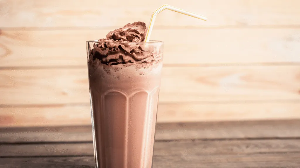 A visually captivating scene of luscious chocolate milkshake variations. In the foreground, a tall glass filled with a rich, creamy chocolate milkshake, topped with swirls of whipped cream and drizzled with dark chocolate syrup. Beside it, an assortment of garnishes and mix-ins, such as crumbled cookies, chopped nuts, and vibrant fresh berries. In the middle ground, a variety of glass bottles and jars containing different syrups, sauces, and toppings, each with a distinct color and texture. The background features a sleek, modern kitchen counter or table, with a minimalist, yet stylish design. Warm, soft lighting creates a cozy, inviting atmosphere, highlighting the shimmering, tempting nature of the milkshake and its customizable elements. Chocolate Shake Recipe Customizations and Creative Variations