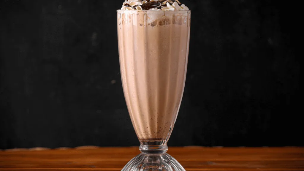 A stainless steel mixing bowl filled with fresh chocolate syrup, whipped cream, and a scoop of rich vanilla ice cream. Beside it, a tall glass, a metal shake mixer, and a selection of toppings including chocolate shavings, crushed Oreos, and a cherry on top. The scene is illuminated by soft, natural lighting, casting warm shadows and highlights that accentuate the creamy textures and glossy surfaces. The overall mood is indulgent and inviting, perfectly capturing the essence of a delectable chocolate milkshake. Chocolate Shake Recipe Ingredients and Essential Tools