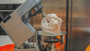 A sunny kitchen countertop, with a large saucepan simmering on the stove. Strands of steam rise from the pan, as golden-brown butterscotch bubbles and caramelizes. Nearby, a wooden spoon stands at the ready, a pat of butter glistening on its surface. The rich, warm scent of brown sugar and vanilla fills the air, inviting the viewer to imagine the decadent taste. In the background, a vintage mixing bowl and whisk hint at the creation of a homemade ice cream base. Soft, natural lighting casts a cozy, inviting glow over the scene, capturing the artisanal process of making this beloved confection.