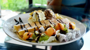 A still life arrangement of various ingredients for homemade butterscotch ice cream, captured in soft, warm lighting. In the foreground, a glass bowl overflows with golden brown sugar, next to a stainless steel measuring cup, a wooden spoon, and a glass jar of vanilla extract. In the middle ground, a ceramic ramekin holds chopped pecans, and a glass pitcher is filled with heavy cream. In the background, a glass bottle of corn syrup and a glass bowl of egg yolks create a cohesive tableau. The composition is arranged on a rustic wooden surface, with a neutral backdrop allowing the ingredients to take center stage.