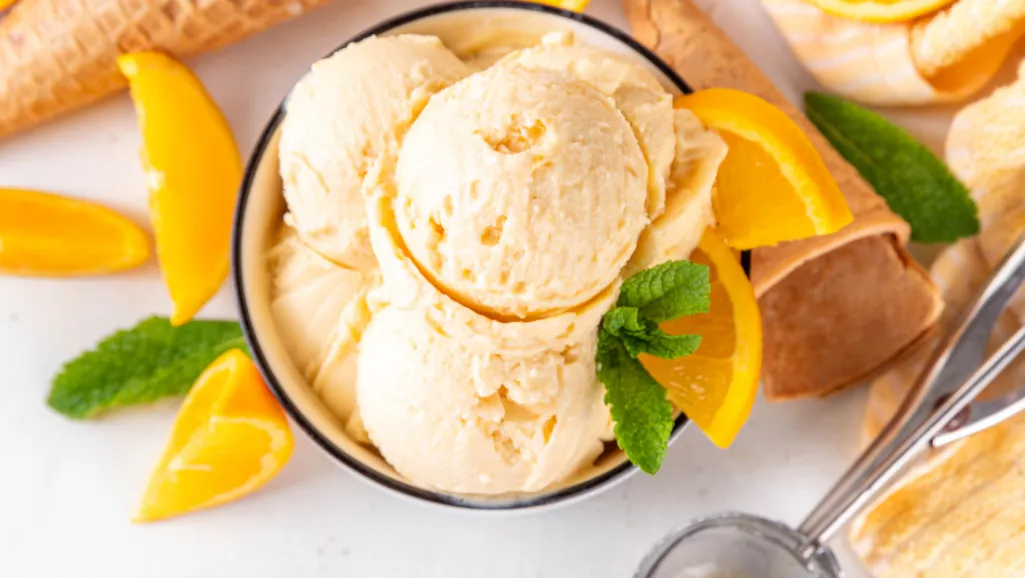A bright, airy kitchen with warm, natural lighting filtering through a window. On a clean, wooden countertop, a stack of freshly-cut orange slices, their juicy segments glistening. Nearby, a vintage hand-crank ice cream maker stands, its metal casing polished to a shine. Scattered around are simple kitchen tools - a juicer, a whisk, a scooper. The scene conveys a sense of homemade, artisanal process, guiding the viewer through the steps of transforming fresh oranges into a delightful, creamy sherbet.