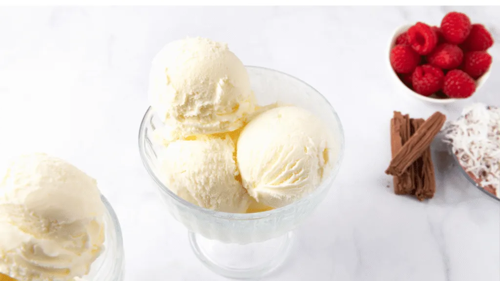 A visually enticing scoop of creamy vanilla ice cream, beautifully presented in a delicate, rustic bowl, sits prominently in the foreground. The ice cream is swirled with subtle hints of yellow and white, glistening with a touch of coconut cream to emphasize its dairy-free nature. In the middle ground, a soft, textured linen napkin adds warmth, while a spoon rests beside the bowl, inviting indulgence. In the background, softly blurred images of fresh vanilla pods and lush green mint leaves create an organic atmosphere, enhancing the natural appeal. The scene is illuminated by warm, diffused sunlight, casting gentle shadows that evoke a cozy, inviting mood. The composition captures the essence of comfort and satisfaction, perfect for a delicious dessert experience. Category winners to match your craving, your dessert, and your budget