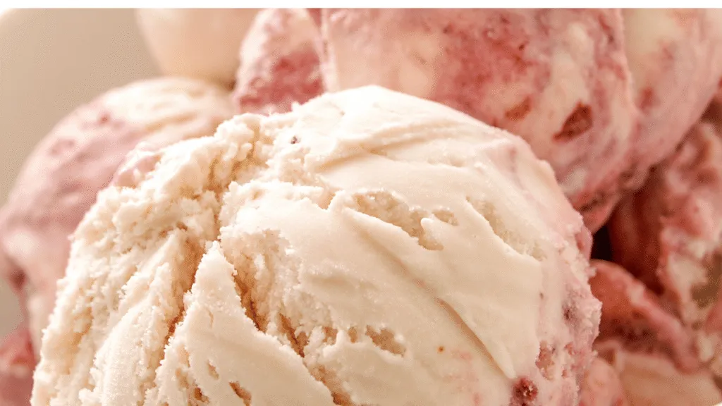 A close-up view of a smooth and creamy vanilla texture, resembling a rich dairy-free ice cream. In the foreground, there are glistening scoops of the ice cream with a delicate sheen under soft, natural lighting that enhances the creamy consistency. The middle layer features a subtle swirl of vanilla bean specks incorporated throughout the ice cream. The background is softly blurred, showcasing a rustic wooden table and hints of fresh vanilla pods and a sprig of mint, creating a warm and inviting atmosphere. The overall mood is comforting and indulgent, evoking the pleasure of enjoying a dairy-free dessert. The image is taken with a macro lens at an angle that captures the inviting texture and lusciousness of the ice cream. How we evaluated these Dairy-Free Ice Cream pints and frozen desserts
