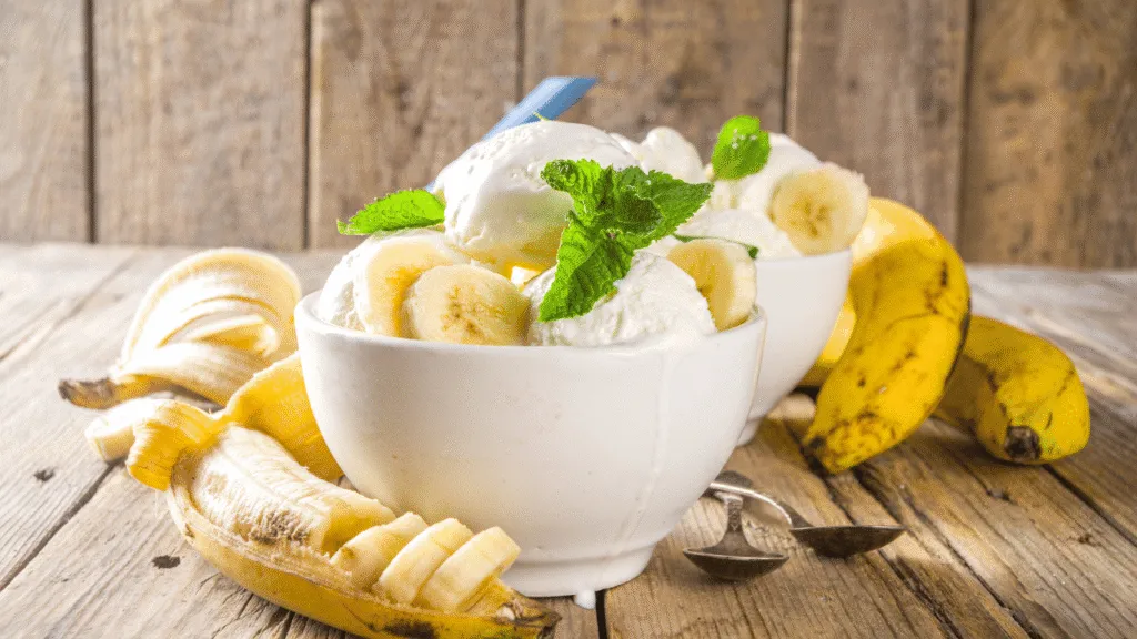 A beautifully arranged composition of ripe, yellow bananas, showcasing their vibrant color and natural sheen. In the foreground, place several whole bananas, some resting on a light wooden cutting board, with a few cut in half to reveal their creamy, soft texture. In the middle, include a small bowl of fresh cream and a scoop of vanilla ice cream, hinting at the ingredients for banana ice cream. The background should be softly blurred, featuring a bright kitchen setting with ingredients like sugar, a measuring cup, and sprigs of mint for freshness. Natural light streams in from a nearby window, creating a warm, inviting atmosphere. Aim for a depth of field that keeps the focus on the bananas and the ice cream while maintaining a cozy, homely vibe. Banana Ice Cream Recipe Basics: Ingredients and Tools