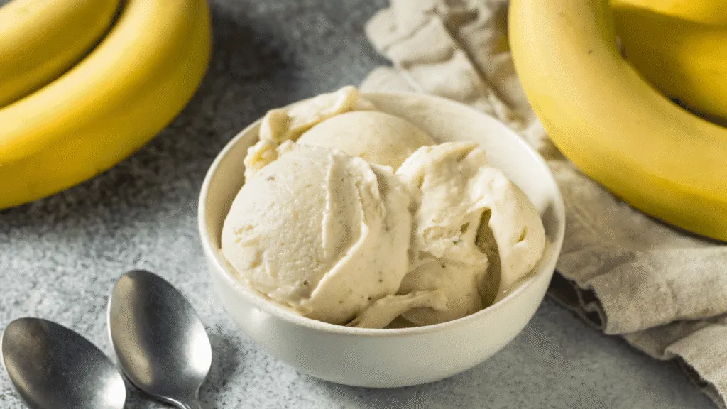 A delicious bowl of peanut butter banana ice cream sits prominently in the foreground, swirled with creamy, rich peanut butter and vibrant yellow banana pieces. The ice cream is garnished with chopped nuts and drizzles of honey, adding texture and sweetness. In the middle ground, a rustic wooden table enhances the homemade vibe, with scattered banana slices and a small jar of peanut butter nearby. The background features a softly blurred kitchen setting with warm, inviting light streaming in, creating a cozy atmosphere. Capture the image with a slight overhead angle, using natural diffused lighting to emphasize the creamy texture and rich colors of the ice cream, making it an enticing focal point that illustrates flavor variations delightfully. Flavor Variations and Mix-Ins for Banana Ice Cream