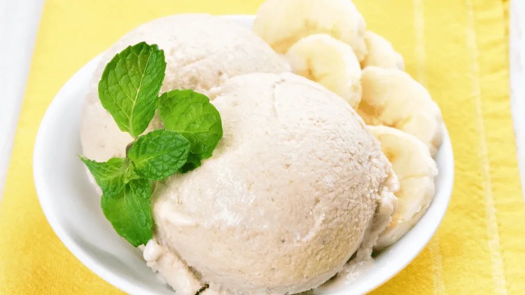 A close-up, vibrant image of several frozen bananas stacked neatly in a clear glass bowl, showcasing their creamy, textured surface. The foreground features a few banana slices artfully arranged beside the bowl, glistening slightly to suggest a fresh chill. In the middle ground, soft-focused kitchen utensils, like a wooden spoon and a mixing bowl, hint at the preparation process. In the background, a sunny kitchen scene is softly illuminated by warm, natural light streaming through a window, creating an inviting atmosphere. The overall mood is fresh and inviting, conveying the promise of a delicious banana ice cream recipe. The photo should be shot at eye level with a shallow depth of field, emphasizing the bananas while gently blurring the background. No text or overlays should be incorporated. How to Prep Frozen Bananas for the Smoothest Texture
