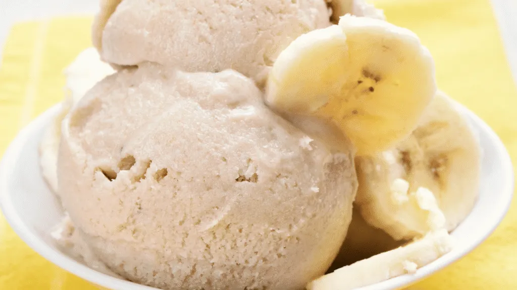 A beautifully styled scoop of homemade banana ice cream sits in a rustic wooden bowl, surrounded by fresh banana slices and a sprinkle of crushed nuts. The ice cream has a creamy, soft texture, reflecting a slight sheen from the light above. In the background, a softly blurred kitchen setting features a touch of vibrant greenery with a potted plant, creating a cozy and inviting atmosphere. The lighting is warm and natural, coming from a window, enhancing the appealing yellow tones of the banana ice cream and ensuring the scene feels fresh and appetizing. The angle captures the scoop from slightly above, focusing on the delightful texture and colors, evoking a feeling of indulgence and freshness. Soft Serve or Scoopable: Freezer Time, Container Tips, and Serving